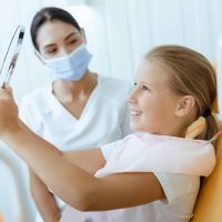 Great dentist work and perfect kids teeth. Happy little girl in medical chair looks in mirror, young female doctor in protective mask and uniform looks at baby in modern clinic at daylight, free space